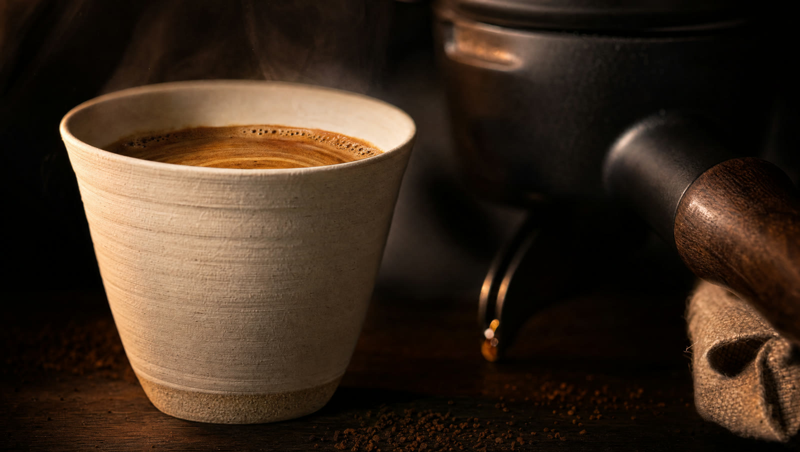 Macro detail of a hand-thrown unglazed ceramic cup of espresso beside a matte blackened steel portafilter with a walnut handle.